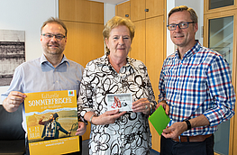 Bürgermeister Otto Steinkamp (rechts) und Frank Jansing (links) bedankten sich mit einer kleinen Aufmerksamkeit bei Renate Bodi, die ihr Motiv „Auf Reisen“ für die Tintenpoeten-Aktion der VarusRegion zur Verfügung stellt.